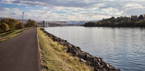 A peaceful riverside path lined with grass and rocks, under a cloudy sky with distant hills and buildings.