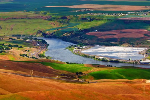 Aerial view of a winding river surrounded by green and brown fields, with a small town in the background.