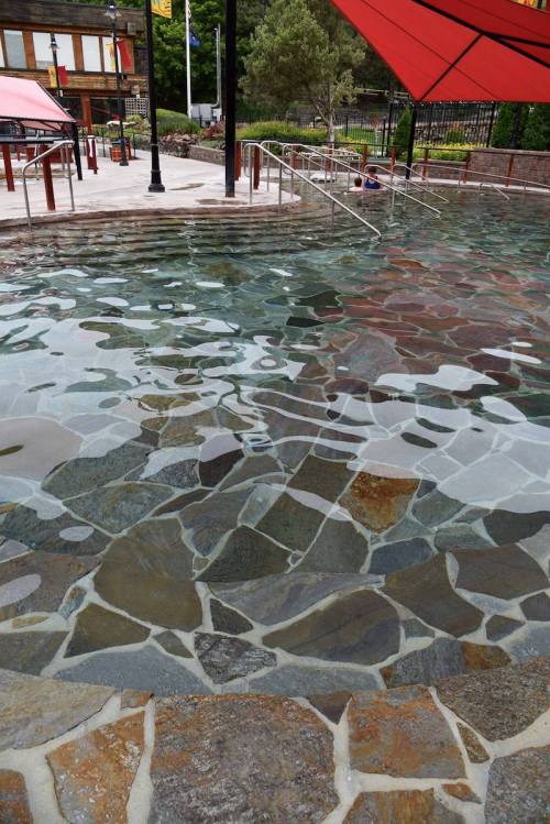 A serene pool with a stone bottom, surrounded by greenery and shaded by a red canopy.