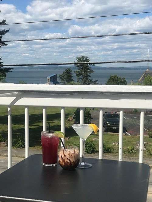 Two cocktails on a table with a scenic view of a lake and cloudy sky in the background.