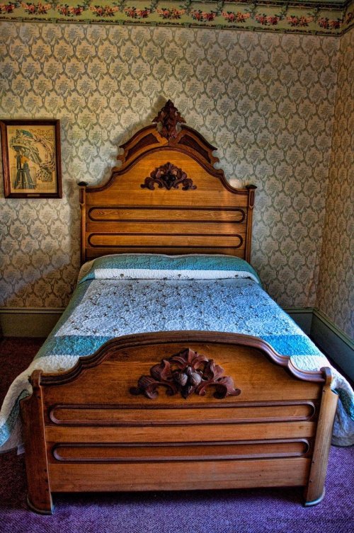 A vintage wooden bed with an ornate headboard and a quilted blanket, set against patterned wallpaper.