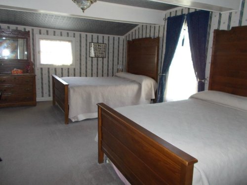 A cozy bedroom with two wooden beds, a dresser, and patterned wallpaper, featuring a window with blue curtains.