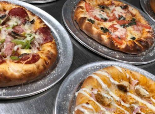 A variety of pizzas on metal trays, featuring toppings like pepperoni, vegetables, and drizzled sauces.