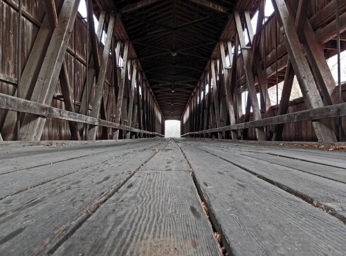 View from inside a wooden covered bridge, showcasing the wooden floor and beams, leading to a bright opening at the end.
