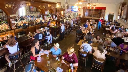A lively bar scene with patrons seated at tables and the bar, enjoying drinks and conversation in a rustic setting.