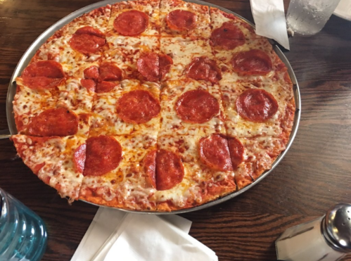 A large pepperoni pizza on a wooden table, sliced into equal pieces, with a glass of water and napkins nearby.
