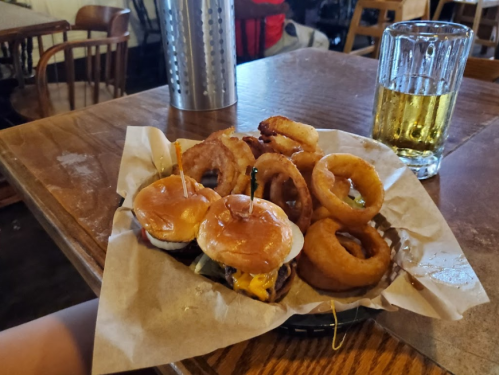 A plate with three mini burgers, onion rings, and a glass of beer on a wooden table in a casual dining setting.