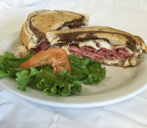 A sliced sandwich with layers of meat and cheese on toasted bread, served with lettuce and a slice of tomato on the side.
