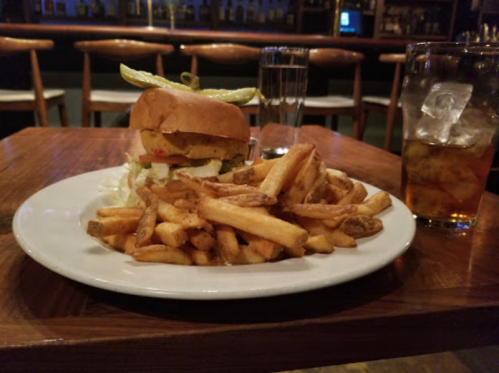 A plate with a burger topped with lettuce and pickles, served with a side of golden fries and drinks in the background.