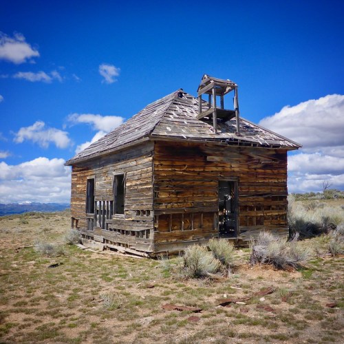 Abandoned wooden house with a damaged roof, surrounded by sparse grass and under a blue sky with clouds.