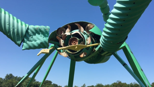A large green sculpture resembling an insect, with spiral wings and a circular body, set against a clear blue sky.