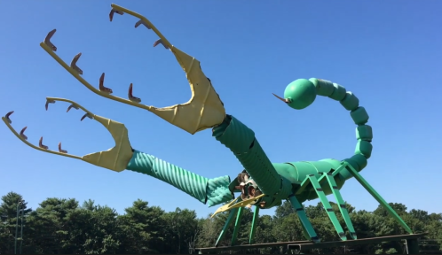 Giant green scorpion sculpture with large claws against a clear blue sky, surrounded by trees.