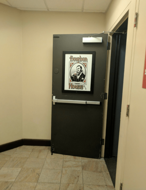 A dark door with a framed poster of a man labeled "Bourbon" next to it, in a corner of a hallway.