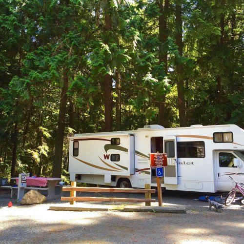 A white RV parked in a wooded campsite, surrounded by tall trees and a picnic table nearby.