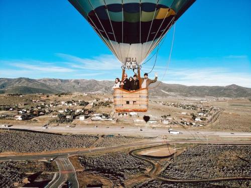 A hot air balloon floats above a scenic landscape with mountains and small towns below, featuring four people in the basket.