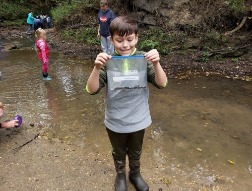 A child in a stream holds a bag of collected specimens, smiling, with other kids playing in the background.