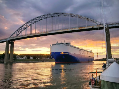 A large cargo ship passes under a bridge at sunset, reflecting on the water.