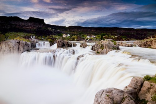 A scenic view of a cascading waterfall surrounded by rocky cliffs and lush greenery under a cloudy sky.