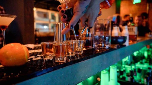 A bartender pours shots into glasses on a bar, with colorful bottles and a lively atmosphere in the background.