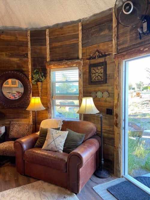 Cozy interior of a rustic room featuring a leather chair, lamps, wooden walls, and a door leading outside.