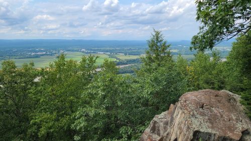 A scenic view from a rocky overlook, featuring lush green trees and a valley with fields under a cloudy sky.