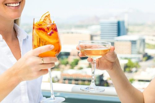 Two people clink glasses of colorful cocktails against a city skyline backdrop.