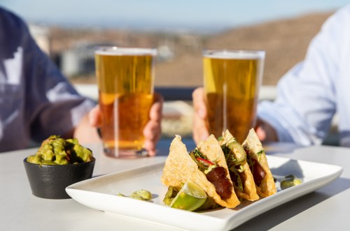 Two glasses of beer are raised in a toast beside a plate of tacos and a bowl of guacamole on a sunny outdoor table.