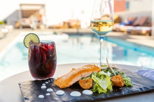 A plate with salmon, salad, and sauce, alongside a glass of white wine and a cocktail, with a pool in the background.