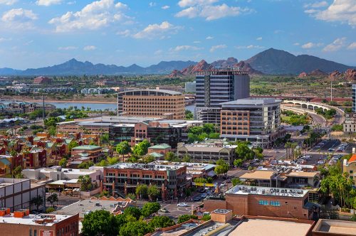 Aerial view of a cityscape with buildings, greenery, and mountains in the background under a partly cloudy sky.