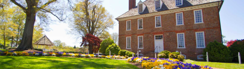 A historic brick house surrounded by colorful flower beds and lush greenery on a sunny day.