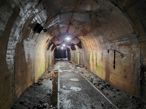A dimly lit underground tunnel with brick walls, a metal grate floor, and patches of moisture on the ground.