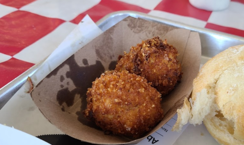 Two golden-brown, crispy fried balls served in a paper tray, with a soft bread roll nearby on a checkered tablecloth.