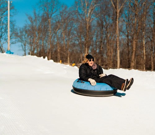 A person in winter attire rides a blue snow tube down a snowy slope, surrounded by bare trees under a clear blue sky.