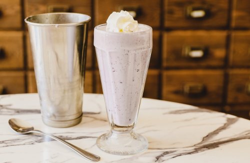 A tall glass of purple milkshake topped with whipped cream, next to a metal shaker, on a marble table.