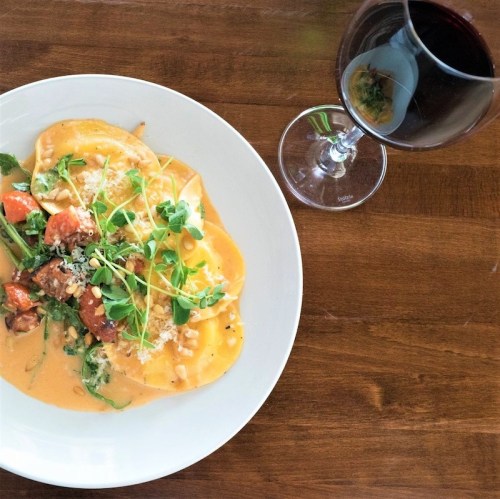 A plate of ravioli with vegetables in a creamy sauce, garnished with herbs, alongside a glass of red wine.