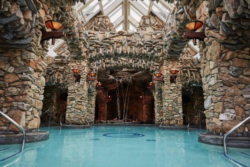 A serene indoor pool surrounded by stone walls and decorative lighting, featuring a waterfall and glass ceiling.