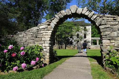 A stone archway frames a path leading to a house, with blooming peonies and greenery on either side.