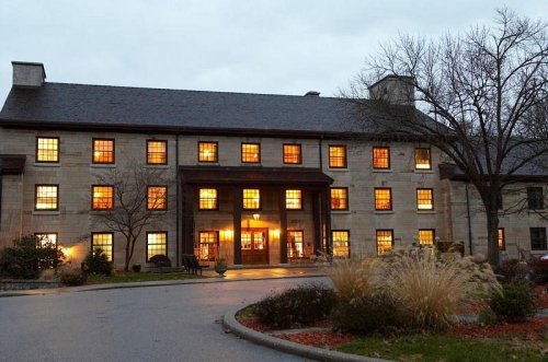 A large, stone building with warm lights glowing from its windows, surrounded by trees and landscaping on a cloudy evening.