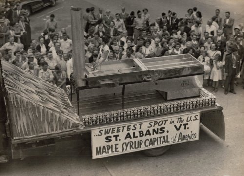 A crowd gathers around a truck displaying a large maple syrup setup, promoting St. Albans, VT as the syrup capital.