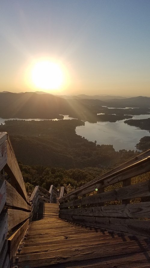 A wooden staircase descends a mountain, with a sunset illuminating a lake and distant hills in the background.