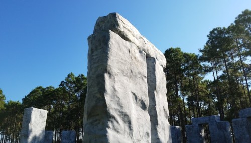 A large stone monolith surrounded by smaller stones, set against a clear blue sky and pine trees.