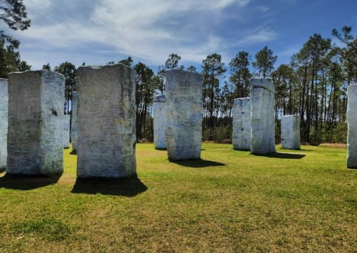 A circle of large, stone-like pillars stands on green grass, surrounded by tall trees and a blue sky.
