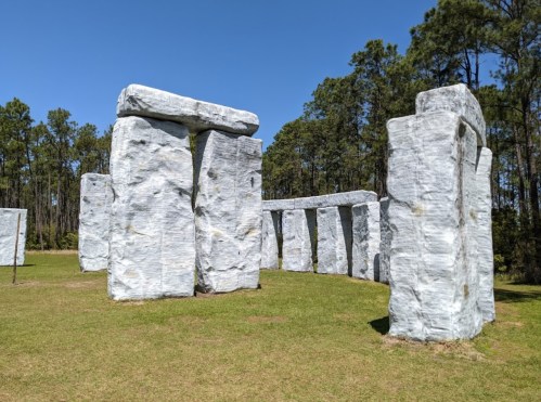 A replica of Stonehenge made of large, gray stone-like structures in a grassy area surrounded by trees.