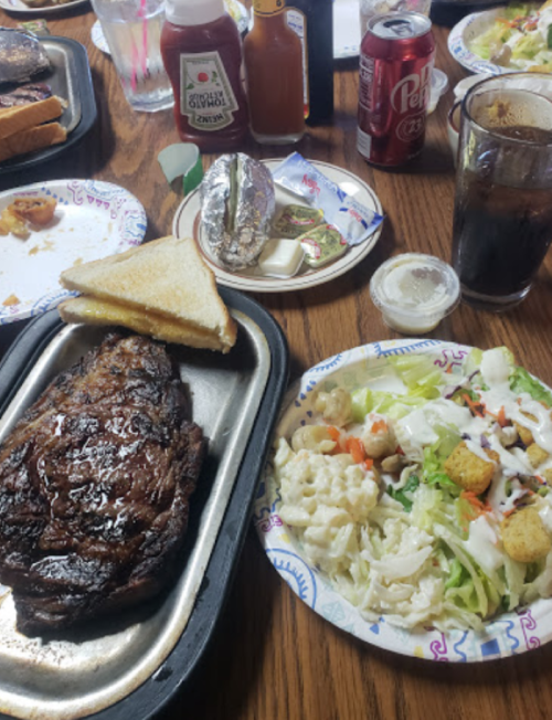 A table spread with a grilled steak, toast, salad, condiments, and drinks, showcasing a hearty meal.