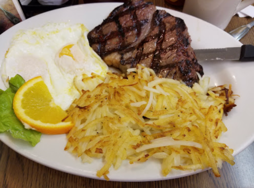 A plate with grilled steak, a sunny-side-up egg, crispy hash browns, and a slice of orange on lettuce.