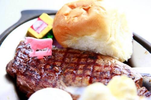 A grilled steak with a pink cow figurine, a soft roll, and butter packets on a plate.