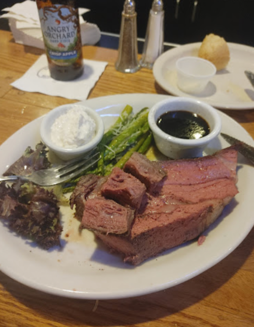 A plate with sliced beef, asparagus, a small bowl of sauce, and a side of sour cream, with a drink in the background.