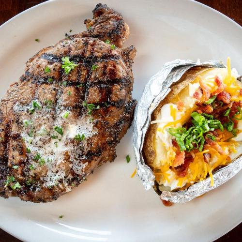 A grilled steak with seasoning, served alongside a baked potato topped with cheese, bacon, and green onions.