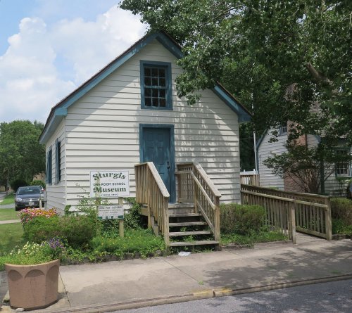 A small, white museum building with a blue door and stairs, surrounded by greenery and a sign reading "Sturgis Museum."