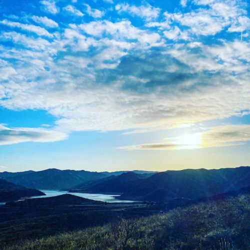 A serene landscape featuring a lake surrounded by hills under a bright sky with scattered clouds at sunset.